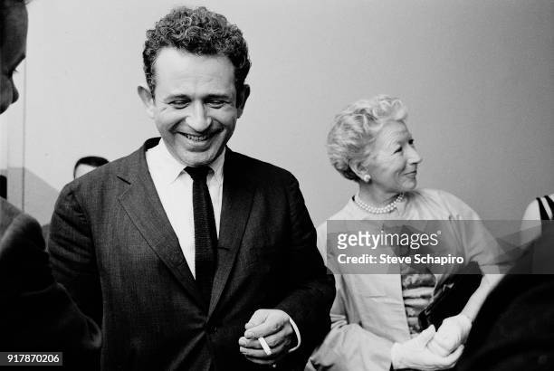 Backstage at Carnegie Hall, American author Norman Mailer laughs, a cigarette in one hand, as behind him, journalist Mary Hemingway talks with...