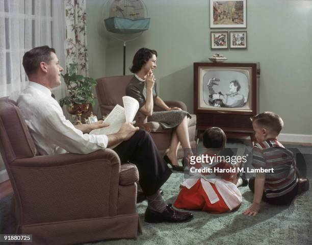 Happy family cheerfully sits in their living room and watches a televisied clown and puppet show, 1957. The father holds an newspaper open to the...