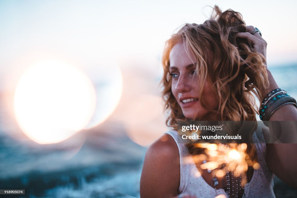 Beautiful boho girl celebrating with sparklers at beach at sunset