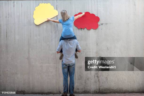 woman sitting on a man's shoulders attaching colourful cloud shapes to concrete wall - wall cloud stock-fotos und bilder