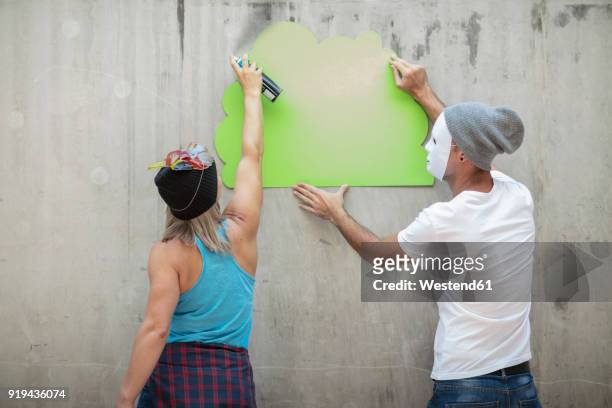 man and woman wearing masks spray painting a concrete wall - hiding behind back stock pictures, royalty-free photos & images