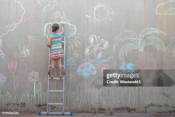 girl standing on ladder drawing colourful pictures with chalk on a concrete wall - chalk wall ストックフォトと画像