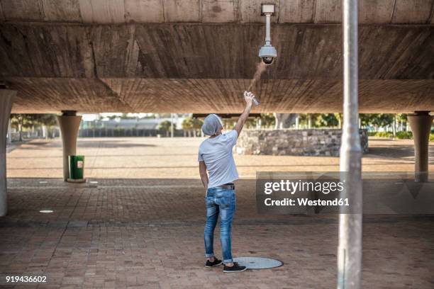 man wearing a white mask spraing a security camera with spraypaint - odd one out obscure stock-fotos und bilder