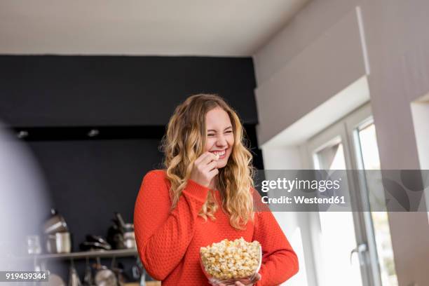 portrait of giggling young woman with bowl of popcorn in the kitchen - snacks stock-fotos und bilder