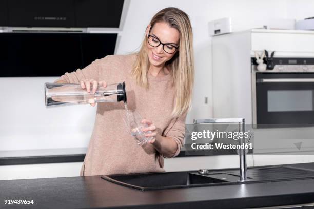 smiling blond woman pouring water into a glass in the kitchen - women with health faucet stock pictures, royalty-free photos & images