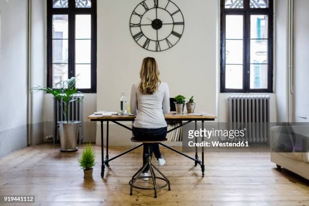 rear view of woman sitting at desk at home under large wall clock - clock person desk stockfoto's en -beelden