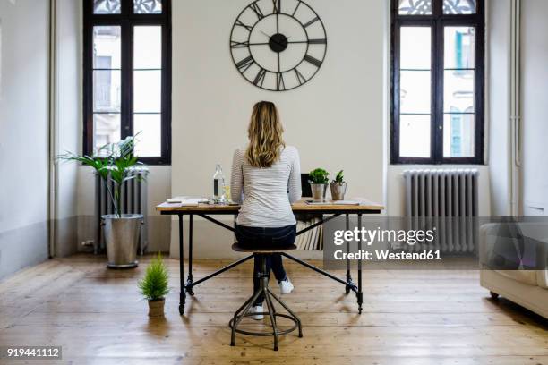 rear view of woman sitting at desk at home under large wall clock - businesswoman under stock pictures, royalty-free photos & images