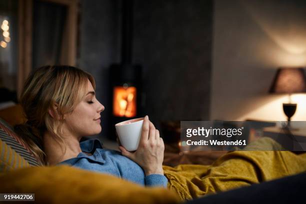smiling woman with cup of coffee relaxing on couch at home in the evening - cosy home stockfoto's en -beelden