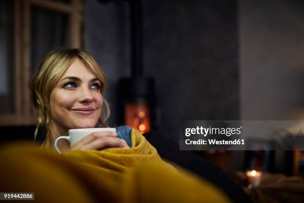 portrait of smiling woman with cup of coffee relaxing on couch at home in the evening - emmitouflé dans une couverture photos et images de collection
