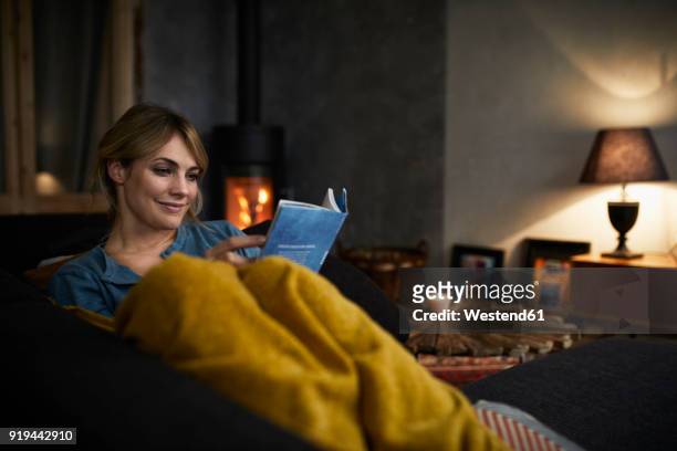 portrait of smiling woman reading a book on couch at home in the evening - reading stock-fotos und bilder