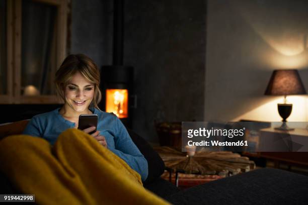 portrait of smiling woman with smartphone relaxing on couch in the evening - cosy stock-fotos und bilder