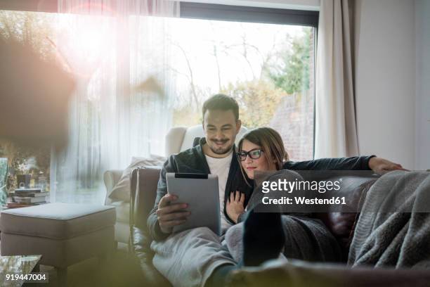 smiling couple lying on couch at home sharing tablet - couple tablet stock-fotos und bilder