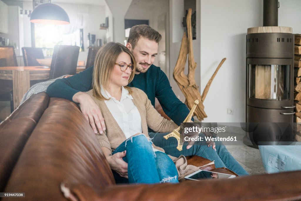 Smiling couple sitting on couch at home with tablet and Eiffel tower model