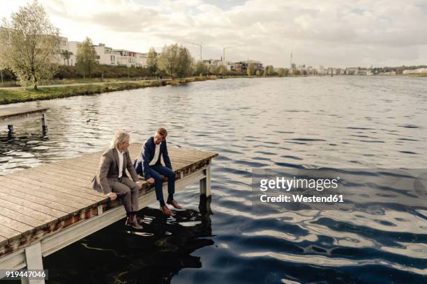 two businessmen sitting on jetty at a lake talking - successor stockfoto's en -beelden