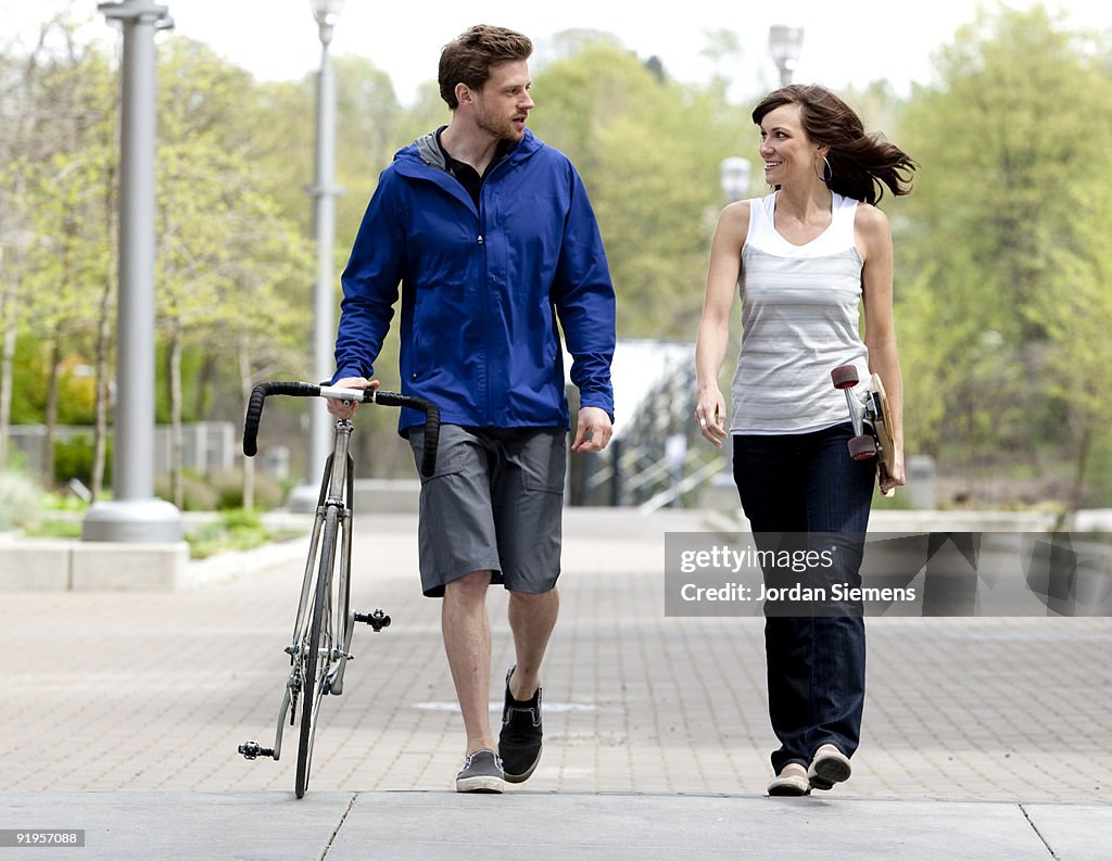 A couple with a bike and a skateboard walk through a city.