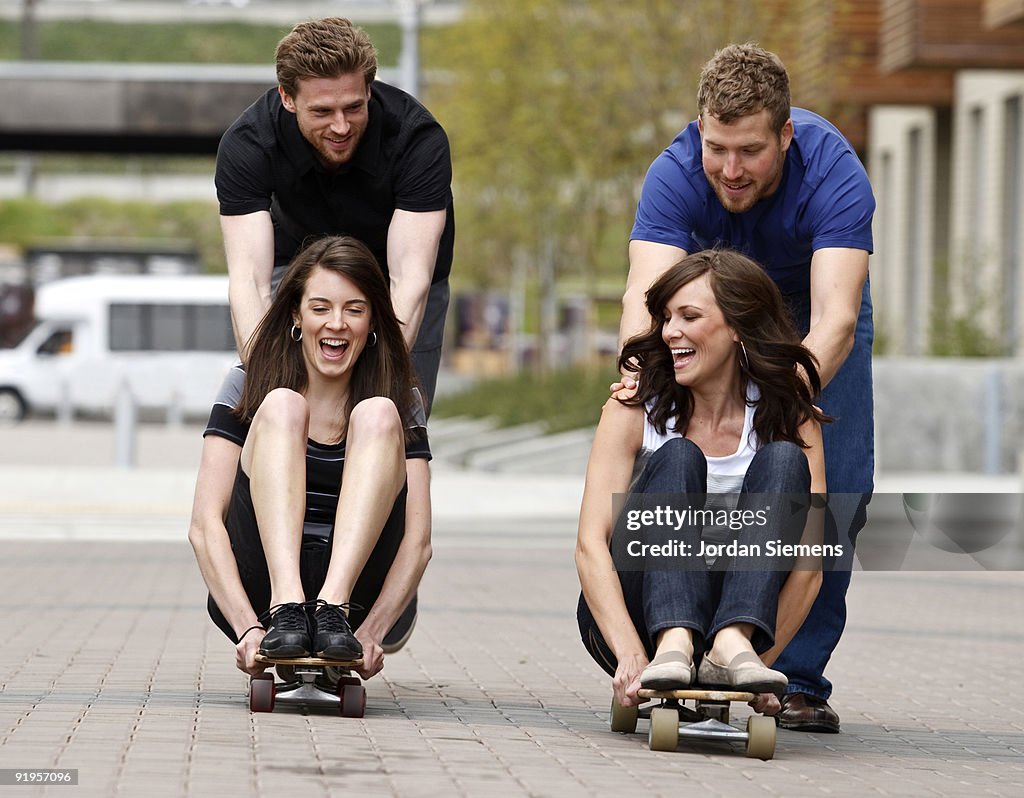 Two young men push two laughing women on skateboards on a city sidewalk.