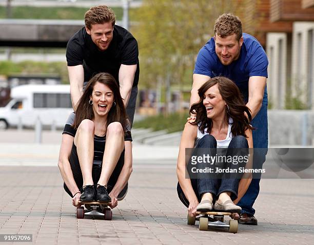 two young men push two laughing women on skateboards on a city sidewalk. - newfriendship stock pictures, royalty-free photos & images