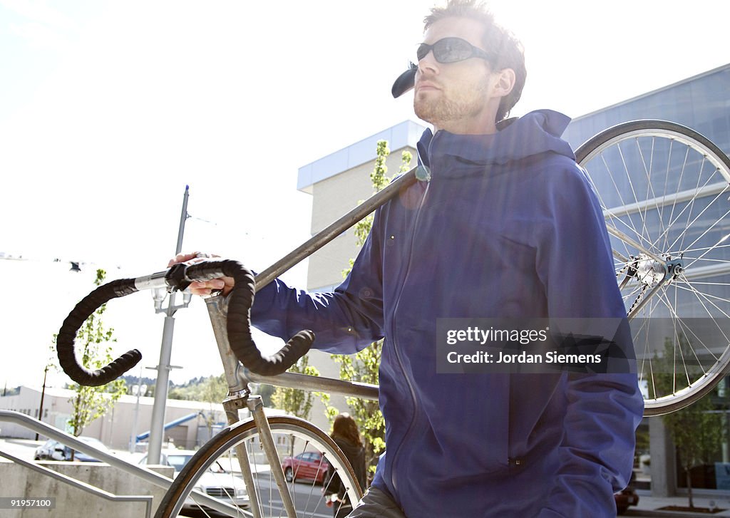 A young bearded man carrying his bicycle on his shoulder in the city. (solar flare)