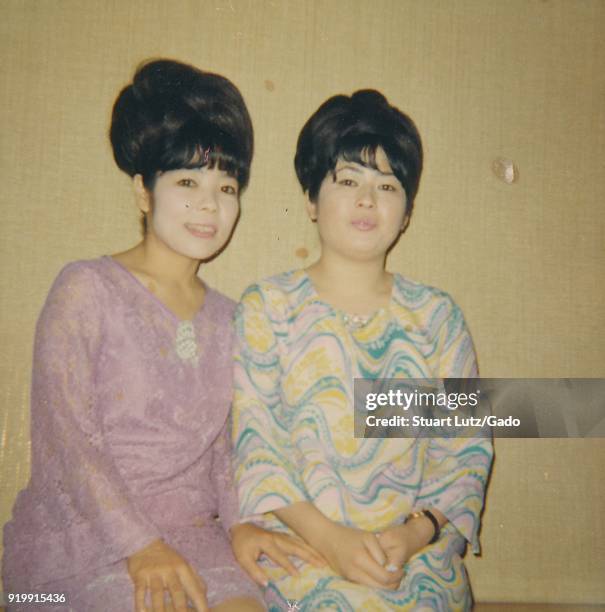 Color photograph, showing a three-quarter view portrait of two Asian women, both wearing colorful dresses, and their hair arranged in high chignons...