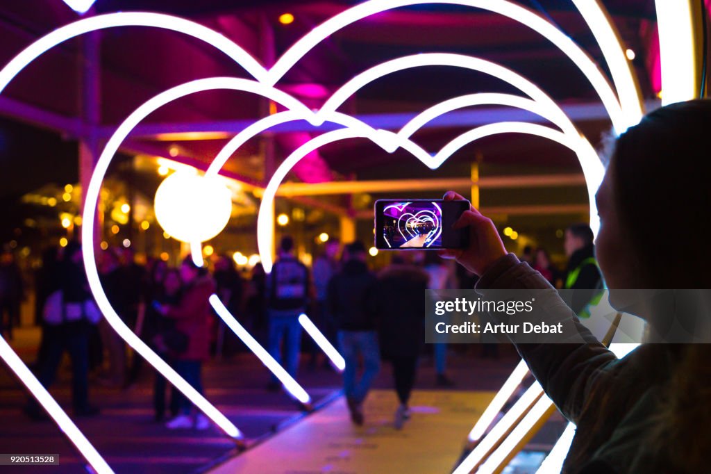 Woman with smartphone taking picture of heart shape lights in the city street.