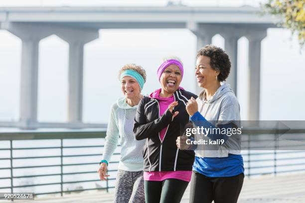 three mature women power walking together on waterfront - active seniors autumn stock pictures, royalty-free photos & images