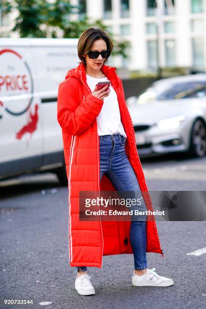 Guest wears sunglasses, a red long puffer coat, a white top, blue denim jeans pants, white sneakers, during London Fashion Week February 2018 on...