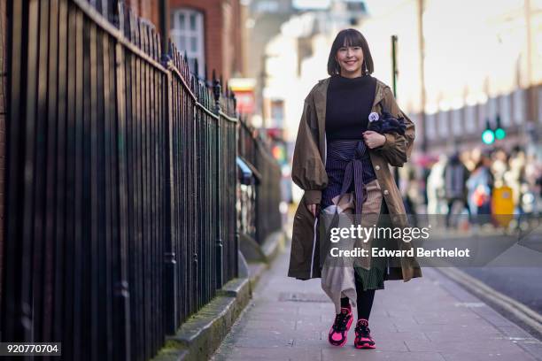 Guest wears a brown trench coat, a black pull over, a striped skirt, pink sneakers shoes, during London Fashion Week February 2018 on February 17,...