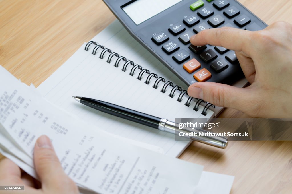Cost calculation or bill payment concept, hand put finger on calculator and black pen on paper notepad with pile of bills in the left hand on wooden table