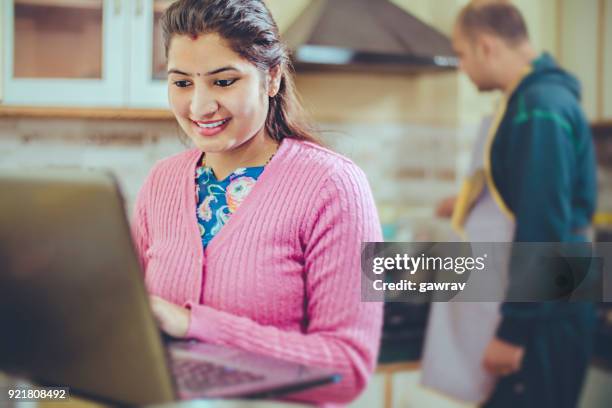 young woman using laptop and husband is cooking food. - working wife stock pictures, royalty-free photos & images