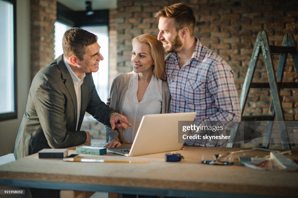 Happy building contractor and young couple communicating while using computer at construction site.