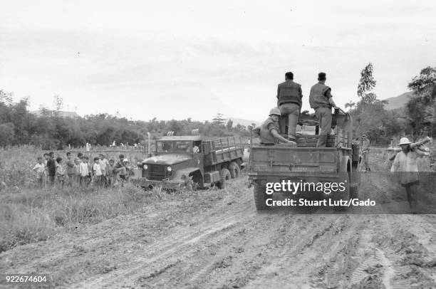 Black and white photograph showing a group of three US soldiers, riding away in the open bed of a pick-up truck, two facing forward, one looking at...