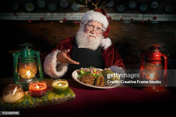 chef santa claus holding a delicious grilled steak dish accompanied with corn and salad in a cozy kitchen counter lit by a candles and oil lamps - tellerlift stock-fotos und bilder