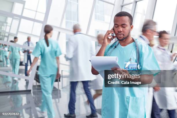 male surgeon with clipboard talking on cell phone in hospital lobby - multitasking nurse stock pictures, royalty-free photos & images