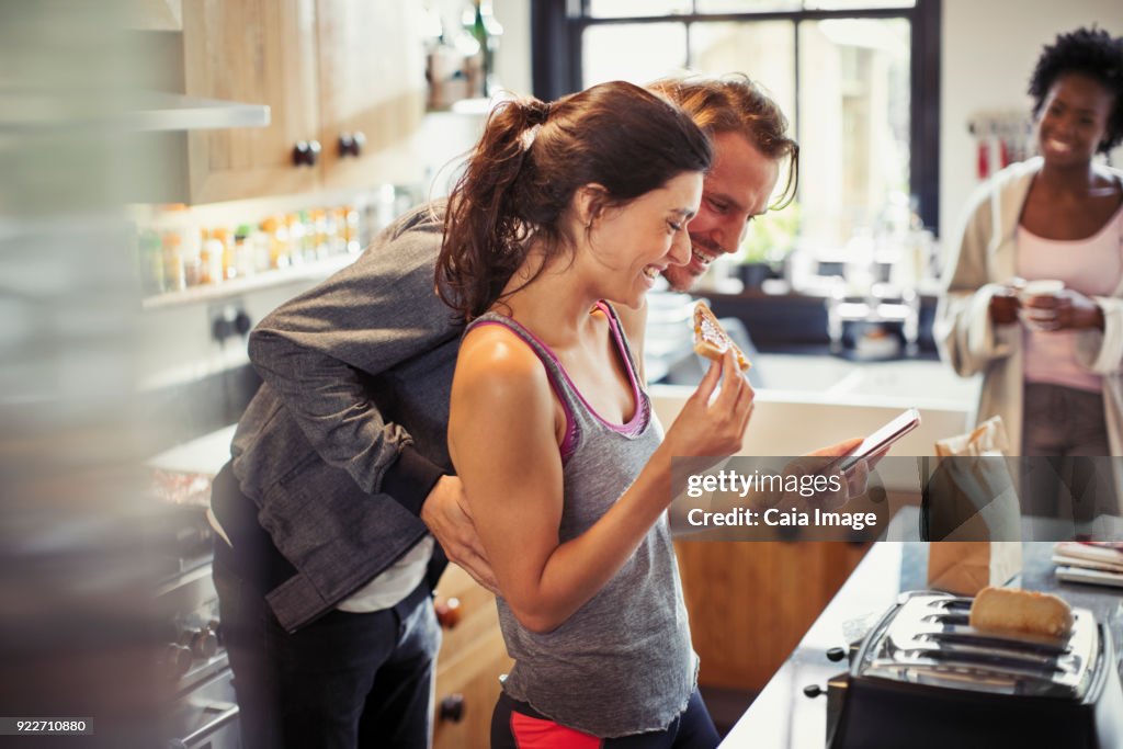 Smiling couple texting with smart phone, eating toast in kitchen