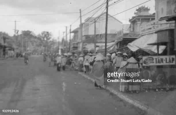 Black and white photograph showing a number of people wearing conical woven hats and walking or standing on the sidewalk of a town or city street,...
