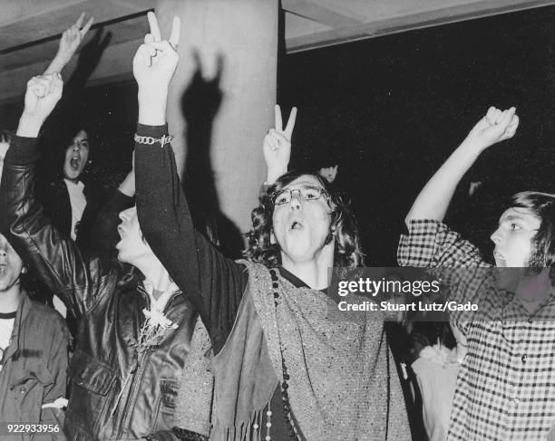 Students wearing hippie attire, including one student with long hair and a poncho, hold their fingers aloft in a peace sign gesture, with some...