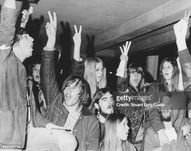 Large group of students wearing hippie attire, including psychedelic patterned clothing, anti-war armbands and long hair, hold their fingers aloft in...