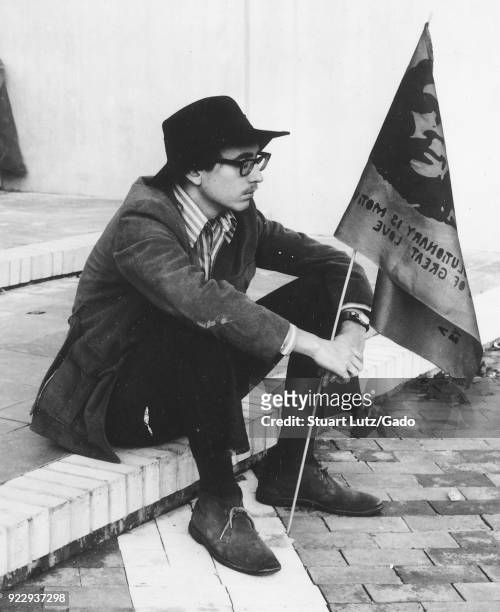 Male student wearing hippie attire, including thick glasses and a fedora hat, sits on a step and holds a flag with the face of revolutionary Che...