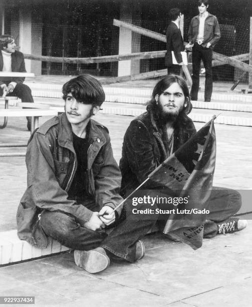 Two male students wearing hippie attire sit on a step, with one holding a flag with the face of revolutionary Che Guevara, during an anti Vietnam War...