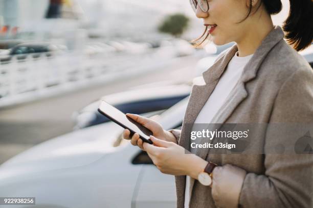 smiling young woman using mobile phone in front of car in city - fachmann auto smartphone stock-fotos und bilder