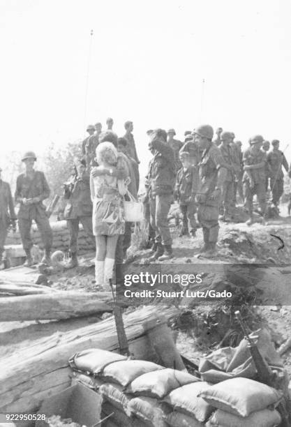 Black and white photograph showing a large group of uniformed US soldiers, one soldier with his arm around a blonde woman who is wearing a patterned...