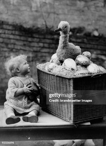 Baby looking unbelievingly at the toy duck sitting on chocolate Easter eggs. Photograph. Around 1935.