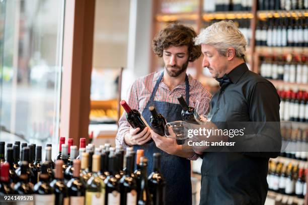 young sales clerk and business owner looking at bottles of wine while owner holds wine glasses at a winery - bottle shop stock pictures, royalty-free photos & images