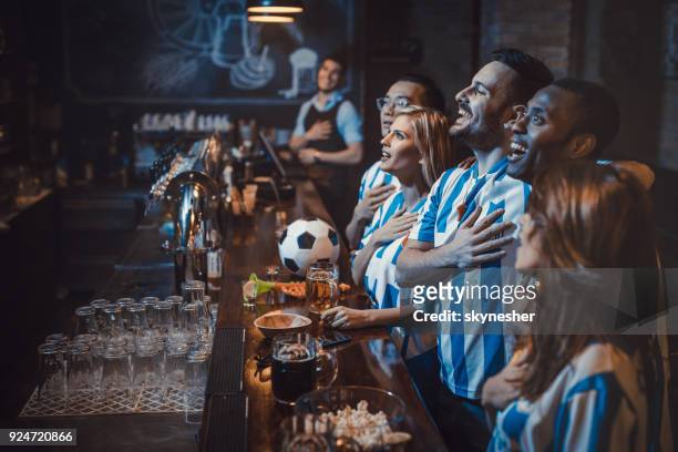 happy soccer fans singing the anthem before the game in a bar. - hands on chest stock pictures, royalty-free photos & images