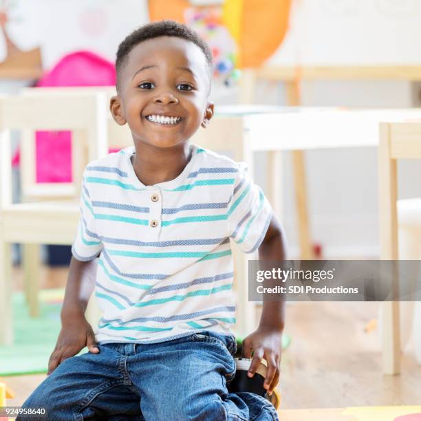 adorable preschool age little boy sits on floor of his classroom and smiles for camera - boy sitting on floor stock pictures, royalty-free photos & images