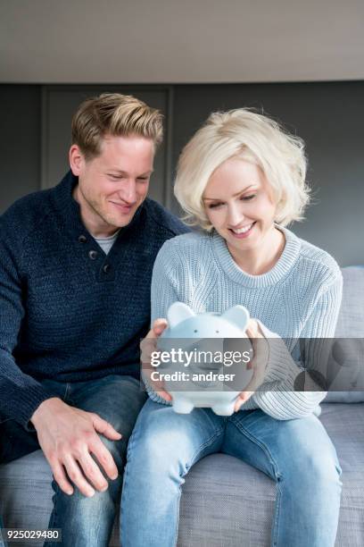 young couple at home both looking at their piggy bank very happy - couple saving piggy bank stock pictures, royalty-free photos & images