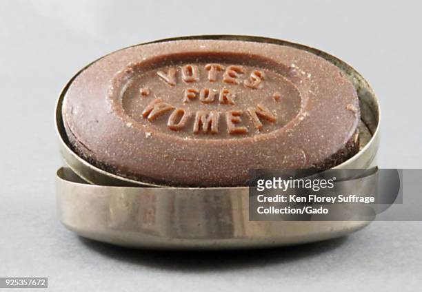 Brown, oval-shaped bar of English glycerin soap, impressed with the words "Votes for Women, " resting in a metal soap container, likely produced for...