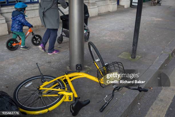 Fallen Ofo dockless hired bike lies on the pavement as a young child scoots past with its mother in Herne Hill, on 26th February 2018, in south...