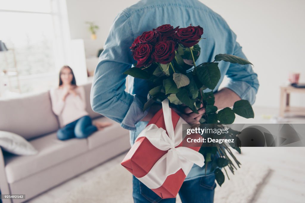 Unexpected moment in routine everyday life! Cropped photo of man's hands hiding holding chic bouquet of red roses and gift with white ribbon behind back, happy woman is on blurred background