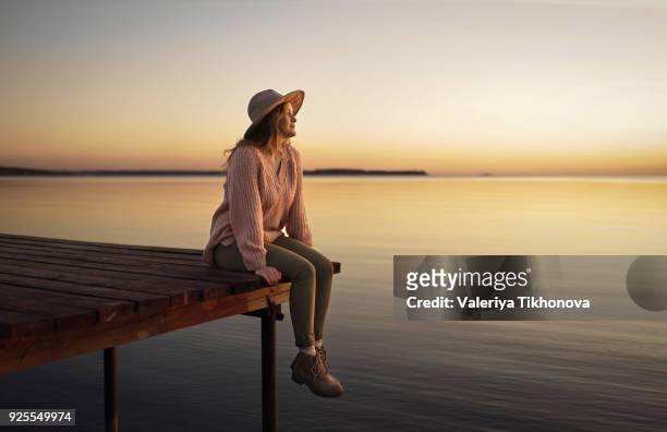 caucasian woman sitting on dock of lake admiring sunset - prier stock-fotos und bilder